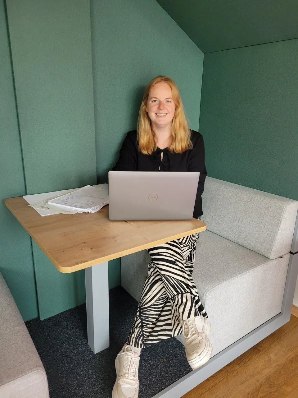 Carina Deing sitzt mit Laptop an einem Holztisch in einer grünen Sitznische.
