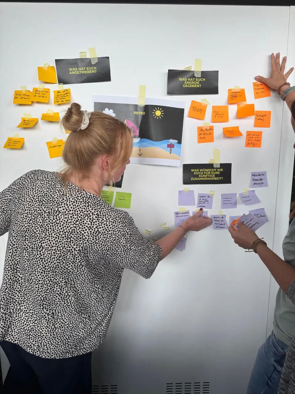 Zwei Teilnehmerinnen kleben Haftnotizen an eine Wand mit Überschriften.
