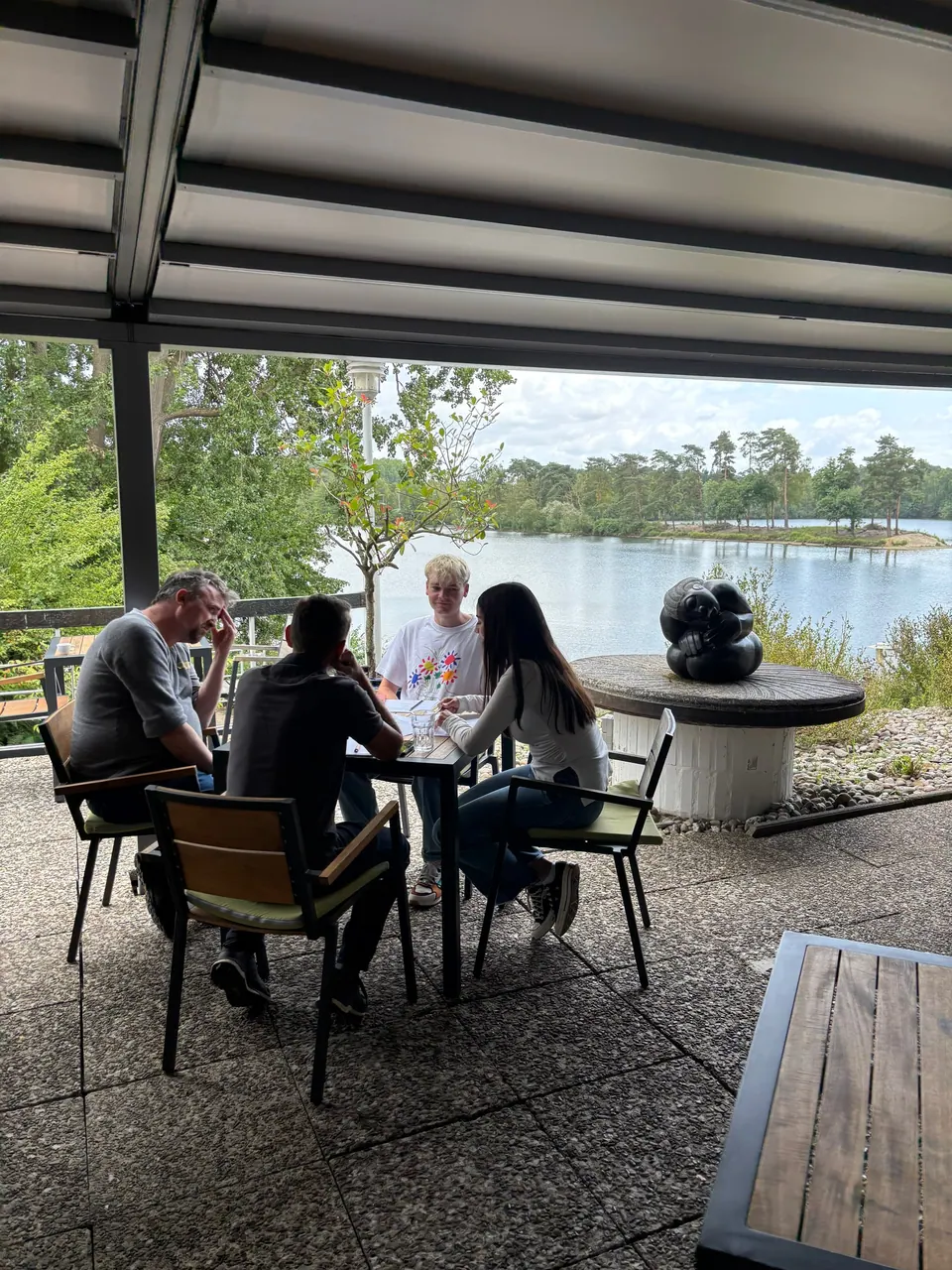 Vier Personen arbeiten auf einer überdachten Terrasse mit Blick auf einen See.