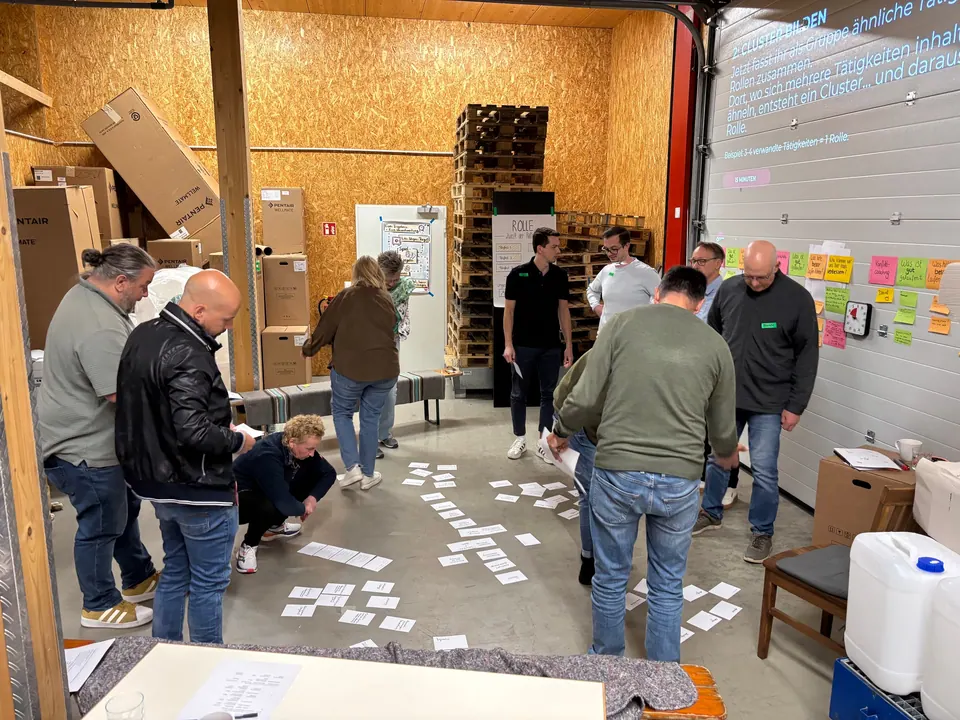 Workshop in einer Halle: Teilnehmende sortieren Karten auf dem Boden, an der Wand hängen bunte Haftnotizen.